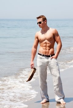 Toned cute handsome male standing on the beach with naked torso showing six pack abs holding shoes in hand wearing sunglasses and hand in pocket