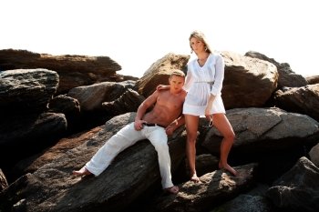 Caucasian guy and girl together on rock formation. Female is wearing long white shirt and holding sunglasses, standing on rocks. Guy showing muscular abs and bare torso wearing beige pants, heaving cool attitude, sitting on rocks. Together hanging out.