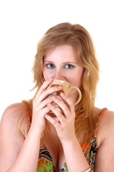A beautiful blond teenager girl looking into the camera and drinking hercoffee from a mug, in an colorful dress over white background.