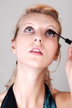 A beautiful blond woman in closeup, doing her makeup and fixing hereyelashes, looking up, for light gray background.