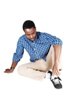 A handsome young African American man in beige pants and a blueshirt sitting on the floor, isolated on white background.
