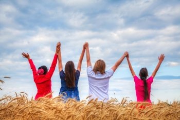 Four young people staying with raised hands at a wheat field at sunset time