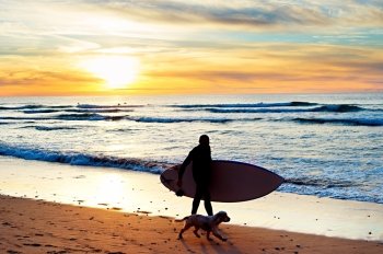 Silhouette of a surfer with a dog walking at sunset on the beach