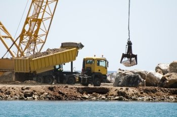 Building a dike. Cranes and excavator put stones in the sea. Trucks carrying stones