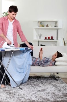 man ironing while his girlfriend is reading a book on the couch