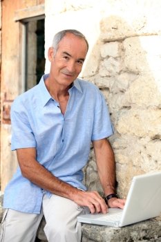 Grey haired man sat by stone wall with laptop