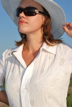The woman in glasses and a hat on a background of the sky. The adult girl in white clothes