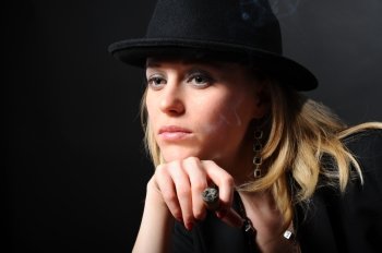 Portrait of the beautiful girl in a hat on a black background with a cigar