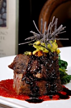 Stacked braised short ribs on a bright red tomato paste sauce.  Garnished with avocado, sliced tortilla chips and a red wine reduction sauce.  Colorful sauteed spinach is displayed behind the meat.