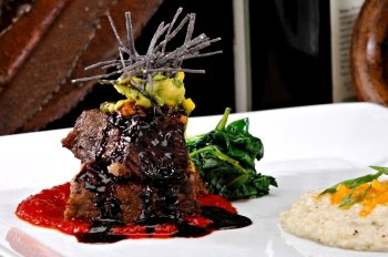 Stacked braised short ribs on a bright red tomato paste sauce.  Garnished with avacado, sliced tortilla chips and a red wine reduction sauce.  Colorful sauteed spinach is displayed behind the meat.
