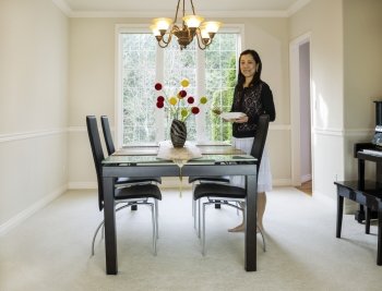 Photo of mature woman placing diner plates and chop sticks  in family formal dining room with daylight coming through large windows in background 