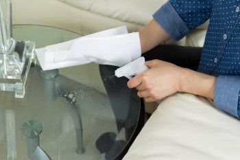 Horizontal photo female hand spraying cleaning solution, from spray bottle, onto dirty glass round end table with paper towels and sofa with partial lamp in background
