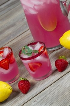 Vertical angled front view of a glass filled with freshly made pink lemonade with whole lemons, strawberries and a partial pitcher in background on rustic wood