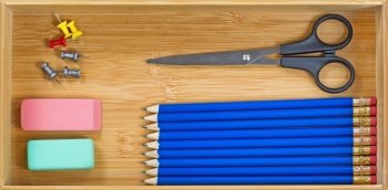 Top view of blue colored pencils, lined up, eraser, scissors, and pin tacks inside wooden desk drawer