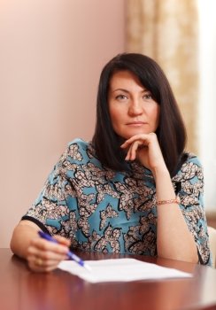Attractive middle-aged woman working at home sitting at a table with a document in front of her and a pen in her hand looking thoughtfully at the camera