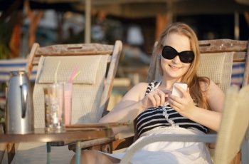 Attractive woman wearing sunglasses relaxing in a deckchair in the sun sending an sms on her mobile phone