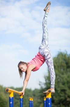Agile attractive young female gymnast balancing on brightly coloured cross bars outdoors in a park with her leg raised high in the air