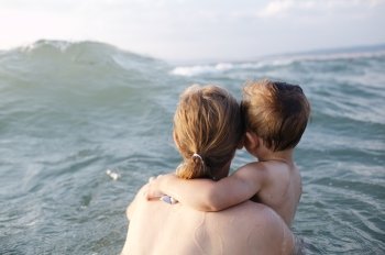 Mother swimming in the sea with her small son in her arms watching an oncoming wave, view from behind them