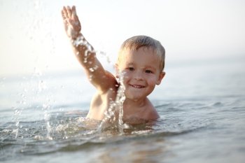 Smiling little boy ducking down in the shallow water at the seaside with a gesture of greeting in a tranquil ocean on a summer day