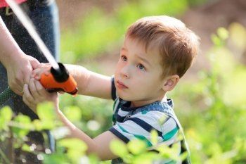 Cute serious little boy playing with a jet of water from a sprinkler being held by his mother in the garden as he stands amongst green leafy shrubs