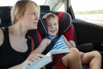 Mother and son traveling by car. Woman reading a book to the kid sitting in child safety seat