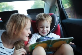 Little boy in a car sitting in child safety seat looking at the pictures in the book, mother watching him