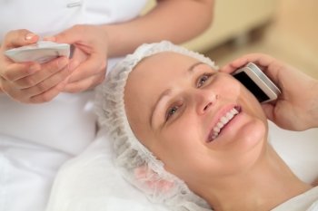 Close-up shot of a young woman talking on the phone at beauty treatment salon while her beautician checking information on cosmetic tube