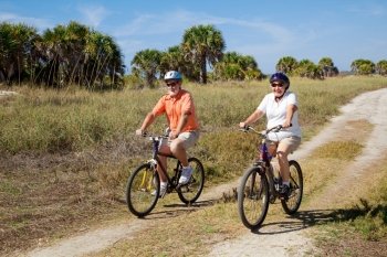 Senior couple bicycling at the beach, wearing safety helmets and sunglasses.
