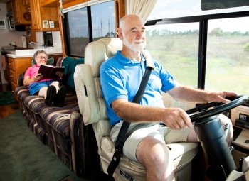 Retired senior couple traveling by motor home.  The husband drives while the wife reads in back.