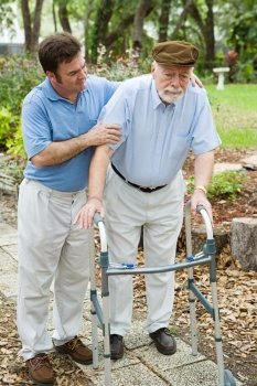 Senior man looks sad as he struggles to walk using a walker.  His adult son is helping him.  