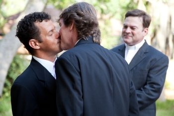 Wedding of handsome gay male couple.  The grooms kiss as the minister looks on.  
