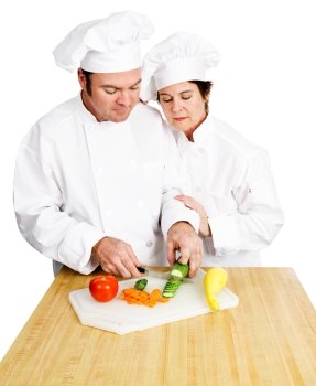 Female chef watches her male student chop vegetables on a cutting board in cooking class.  Both are in full chef’s whites.  Isolated on white background.