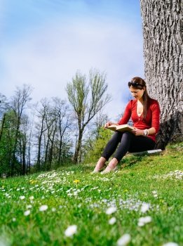 Photo of a beautiful young woman reading a book sitting against a tree in early spring.