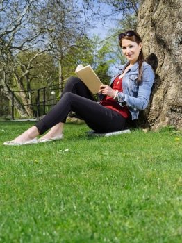 Photo of a beautiful young woman reading a book sitting against a tree in early spring.