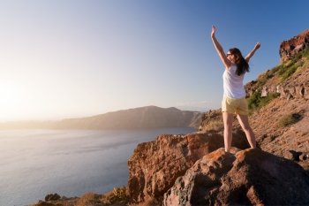 Happy woman on the rock with hands up. Santorini island, Greece, view on Caldera and Aegean sea at sunset. Winner, success, active, travel concepts