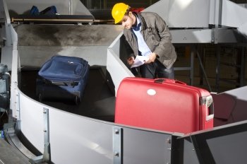 An airport official checking luggage on a conveyor belt, wearing a hard hat and earplugs