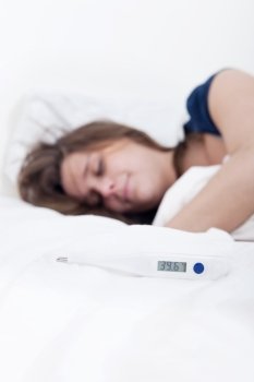 Thermometer on the duvet indicating 39,6 degrees Celcius, with Sick young woman lying in bed with high fever, out of focus, in the background