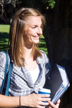 Young female student, holding a dossier with notes and a cup of coffee, smiling in the sunlight