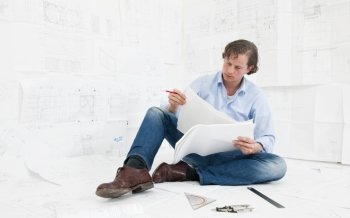 Young mechanical engineer, surrounded by huge technical drawings, sitting on the floor on more designs, going over the technical details, with a red pencil in his hand
