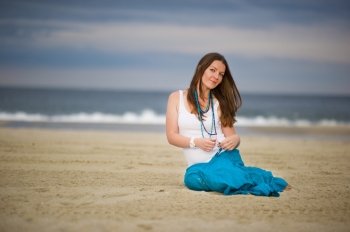 Young beautiful woman in white cami and turquoise skirt sits on sand near the ocean