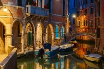 Lateral canal and pedestrian bridge in Venice at night with street light illuminating bridge and houses, with docked boats, Italy