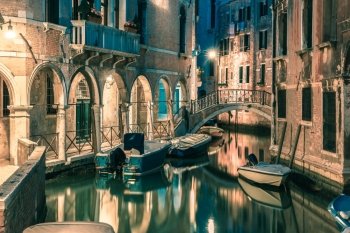 Lateral canal and pedestrian bridge in Venice at night with street light illuminating bridge and houses, with docked boats, Italy. Toning in cool tones