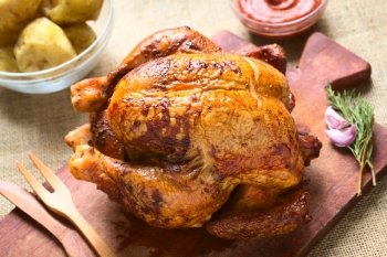 Roast chicken on wooden board with potatoes and ketchup, photographed with natural light (Selective Focus, Focus on the front of the chicken)
