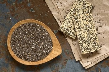 Chia seeds (lat. Salvia hispanica) on small bamboo plate with chia-sesame-honey granola bar on the side, photographed overhead on slate with natural light. Chia seeds are considered a superfood containing protein, omega fat, minerals, antioxidants.