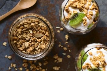 Crunchy almond and oatmeal granola in jar with yogurt kiwi granola parfait in glasses on the side, photographed overhead on slate with natural light (Selective Focus, Focus on the top of the granola and the parfaits)