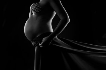 Pregnant woman in silk veil demonstrating her belly.  Black and white studio shot on black background