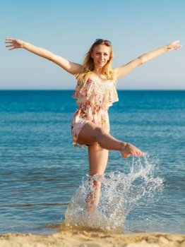 Vacation. Girl in summer dress splashing water on the coast. Young woman having fun relaxing on the sea. Summertime.