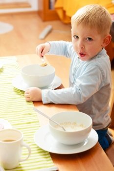 Happy childhood. Blond boy kid child eating corn flakes cereal with milk breakfast morning meal at the table. Home.