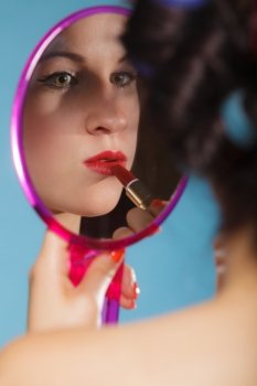 young woman preparing to party, getting ready for going out. Girl styling hair with curlers applying make up red lipstick looking at mirror retro style blue background