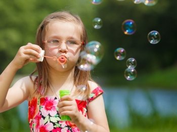 Children happiness and carefree concept. Little girl having fun blowing soap bubbles in park green blurred background